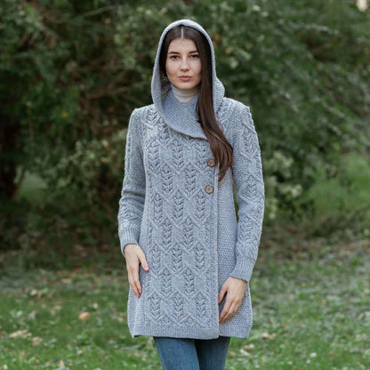 A woman weaing a Ladies Aran Leaf Cable Knit Coat, hood up, standing in front of a green tree