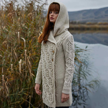 A woman wearing a parsnip-colored Ladies Aran Leaf Cable Knit Coat cardigan stands outdoors near tall reeds and water, with blurred hills in the background.