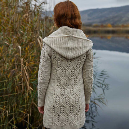 A woman viewed from behind wearing a Ladies Aran Leaf Cable Knit Coat cardigan in a light oatmeal beige color, standing by a calm lake with reeds and mountains in the background.