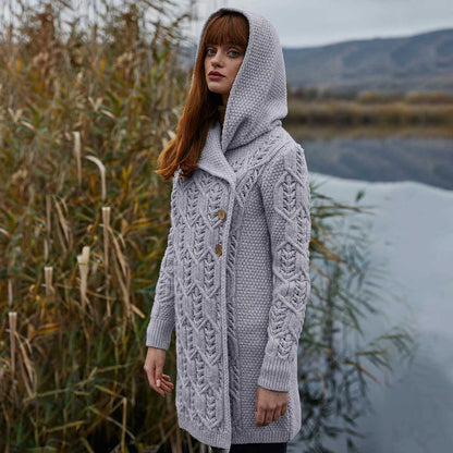 A woman wearing a gray Ladies Aran Leaf Cable Knit Coat hooded cardigan coat stands outdoors near tall reeds and water, with blurred hills in the background.