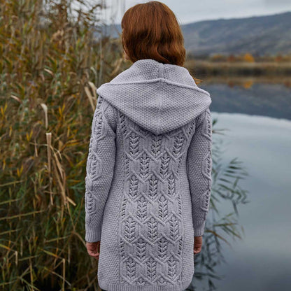 A woman wearing a gray Ladies Aran Leaf Cable Knit Coat by a body of water with mountains in the background.