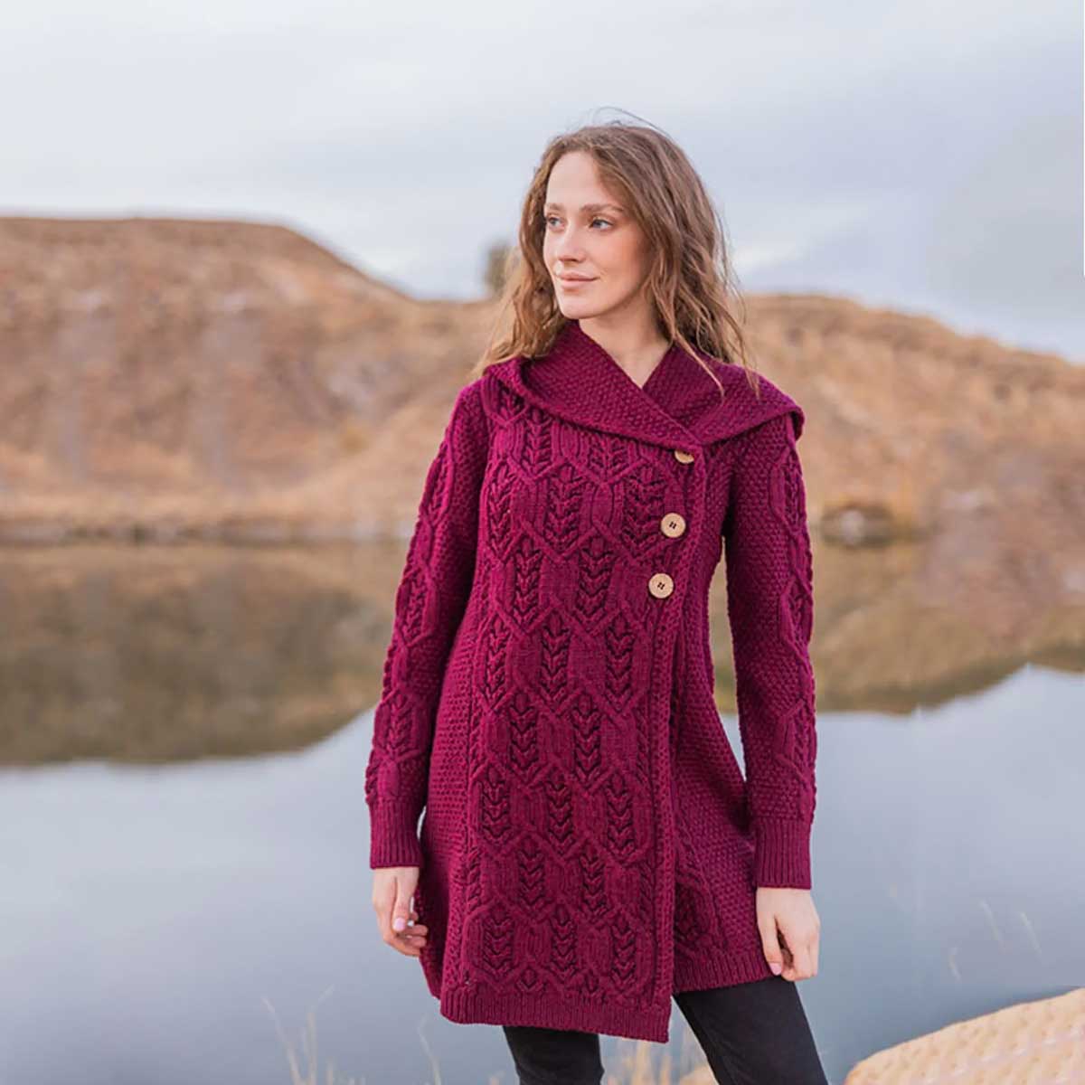 a woman wearing a wine-colored ladies aran leaf cable knit coat with wooden buttons, standing outdoors in front of a calm lake and rocky hills.