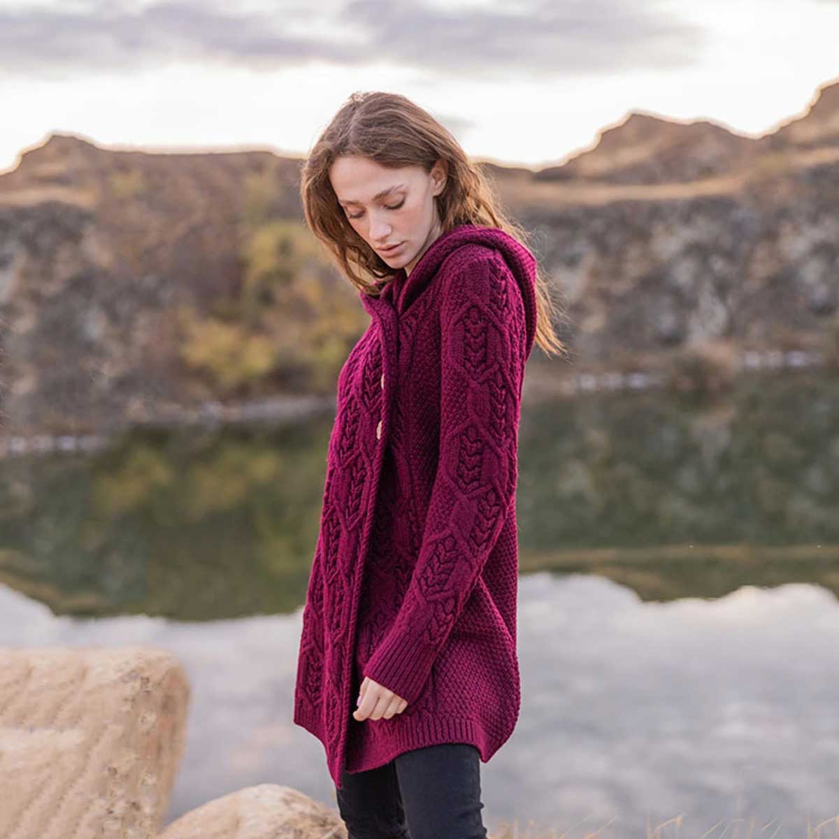 a woman wearing a wine-colored ladies aran leaf cable knit coat stands outdoors near a lake with mountains in the background.