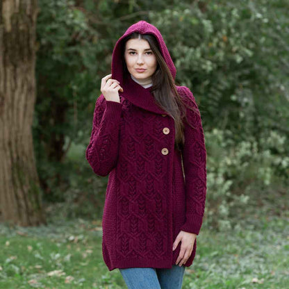 A woman wearing a wine-colored Ladies Aran Leaf Cable Knit Coat with wooden buttons, hood up, in an outdoor setting.