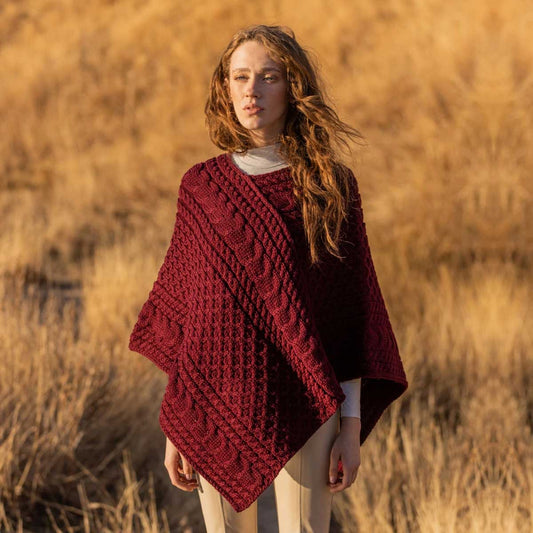 A woman standing outdoors in a field of dry golden grass, wearing a Ladies Honeycomb Cable Knit Poncho in wine color, over a light top and cream pants.