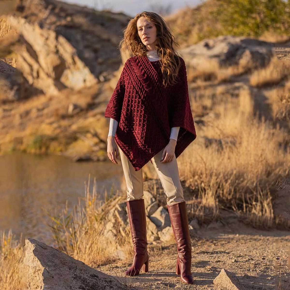 a woman stands outdoors in a rocky, dry landscape wearing a wine colored ladies honeycomb cable knit poncho, beige pants, and burgundy knee-high boots.