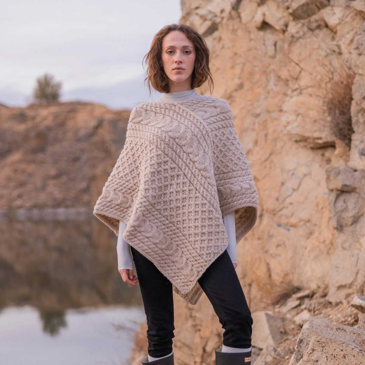 a woman wearing a ladies honeycomb cable knit poncho in color parsnip, styled over a white long-sleeve top and black pants, standing outdoors against a rocky cliffside background.