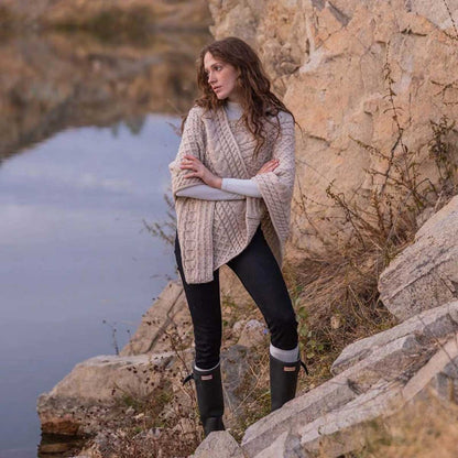 A woman stands outdoors on rocky terrain beside a body of water, wearing a Ladies Honeycomb Cable Knit Poncho in color parsnip, black pants, and dark rain boots.