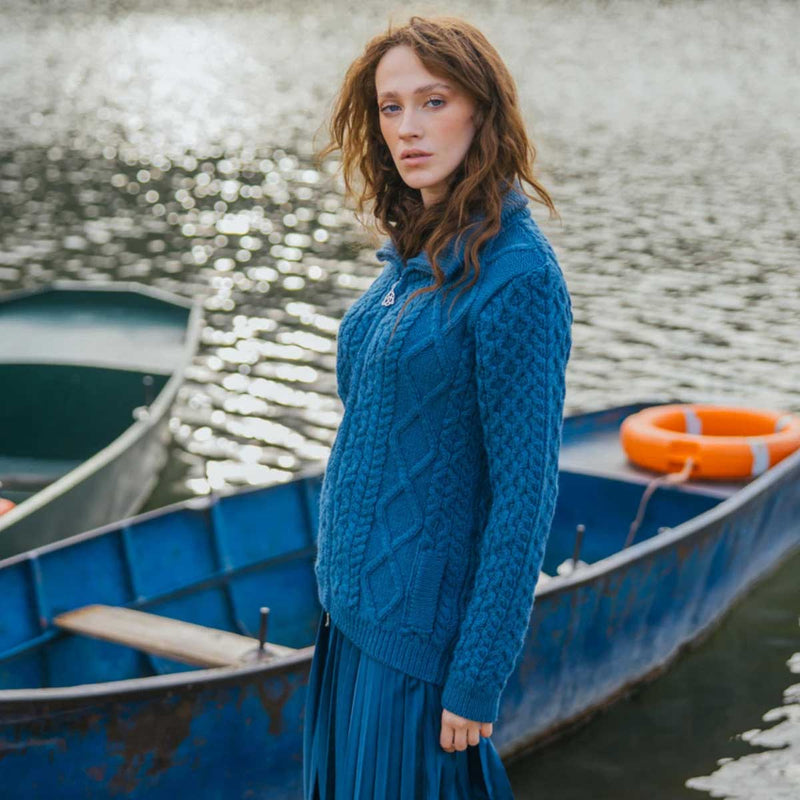 A woman stands in a blue rowboat on a lake, wearing an Teal Ladies Cable Knit Bomber Jacket and blue skirt. Another blue boat and an orange life preserver are visible in the background.