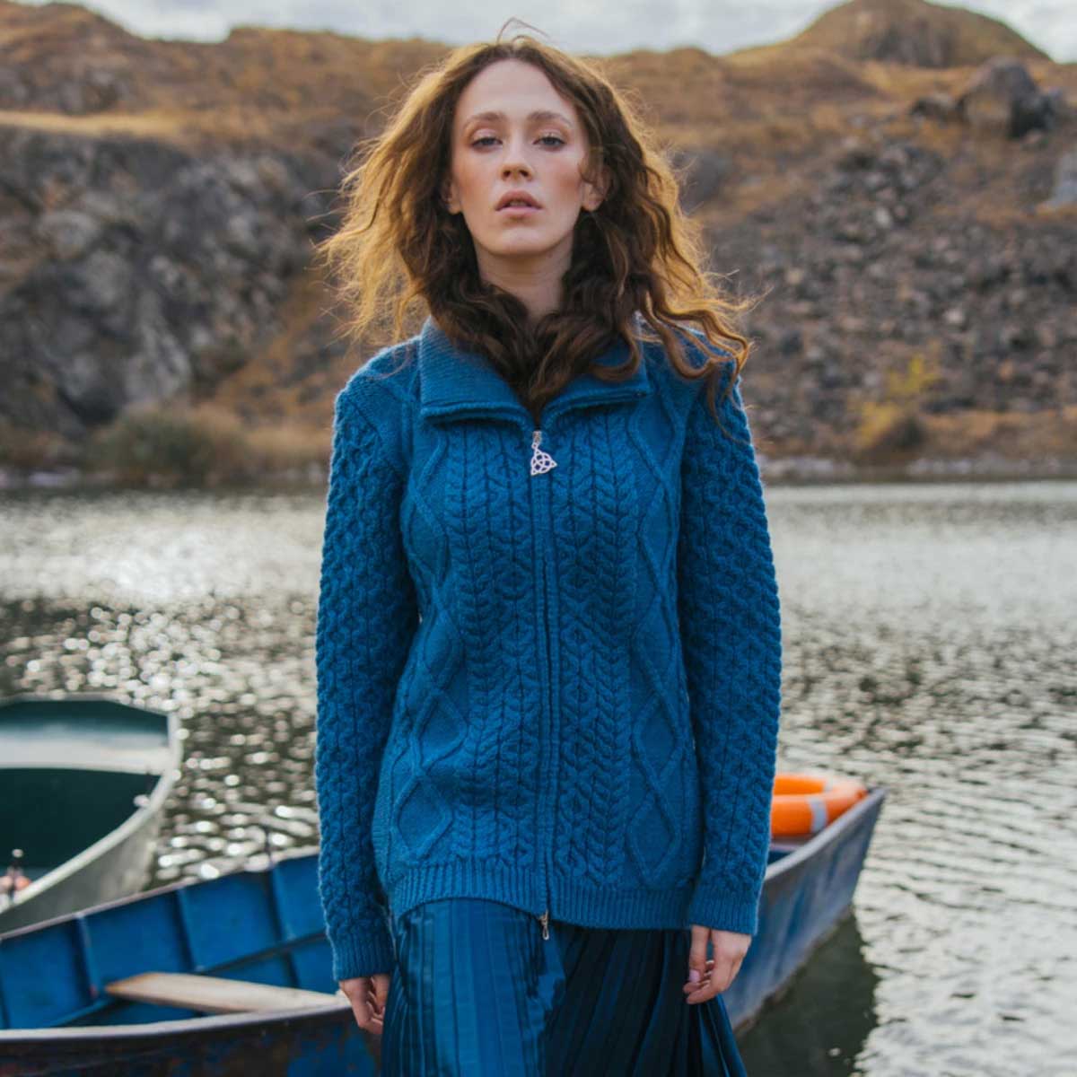 a woman stands by a lake wearing an teal ladies cable knit bomber jacket and blue skirt, with boats and a rocky shoreline in the background.