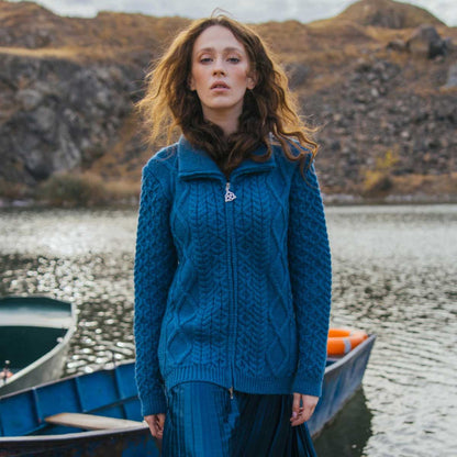 A woman stands by a lake wearing an Teal Ladies Cable Knit Bomber Jacket and blue skirt, with boats and a rocky shoreline in the background.