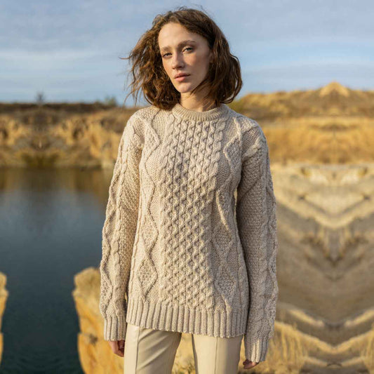 Woman wearing a parsnip colored Fisherman Cable Knit Ladies Sweater standing by a body of water with a natural landscape in the background.