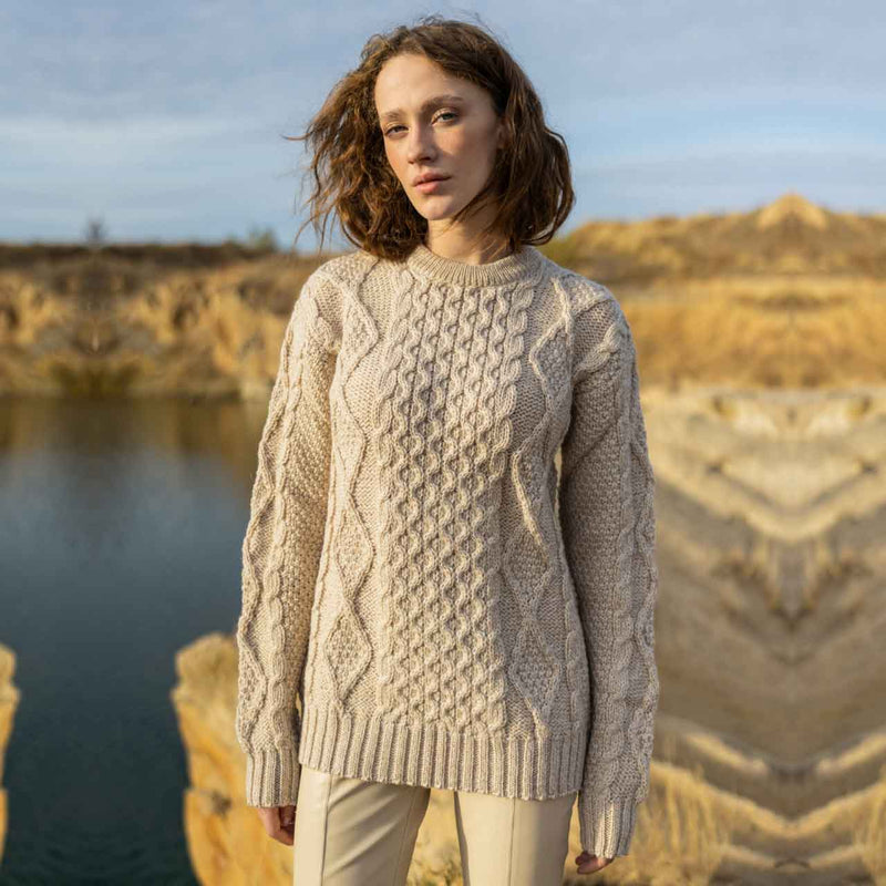 Woman wearing a parsnip colored Fisherman Cable Knit Ladies Sweater standing by a body of water with a natural landscape in the background.