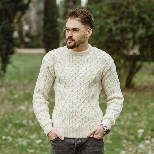 Man wearing a natural colored Fisherman Cable Knit Mens Sweater standing outdoors with greenery in the background