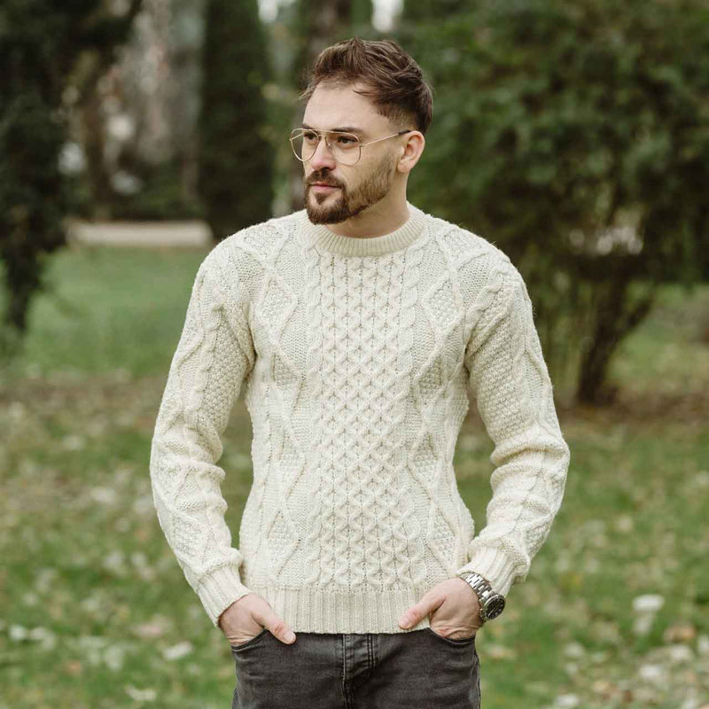Man wearing a natural colored Fisherman Cable Knit Mens Sweater standing outdoors with greenery in the background