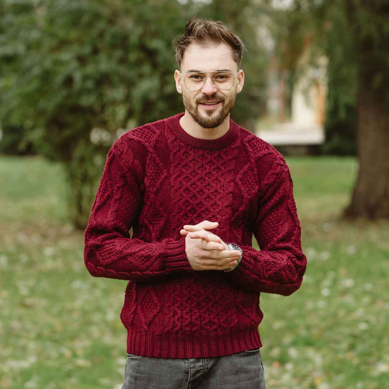man wearing a wine colored fisherman cable knit mens sweater standing outdoors with greenery in the background