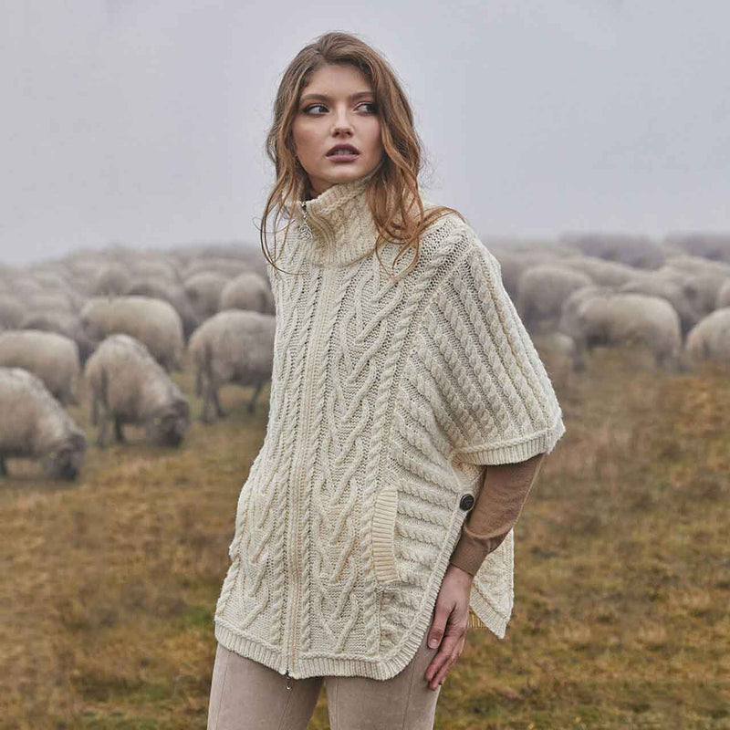 Woman wearing an Aran Knit Funnel Neck Poncho Jacket, in color parsnip, standing in a field with sheep.