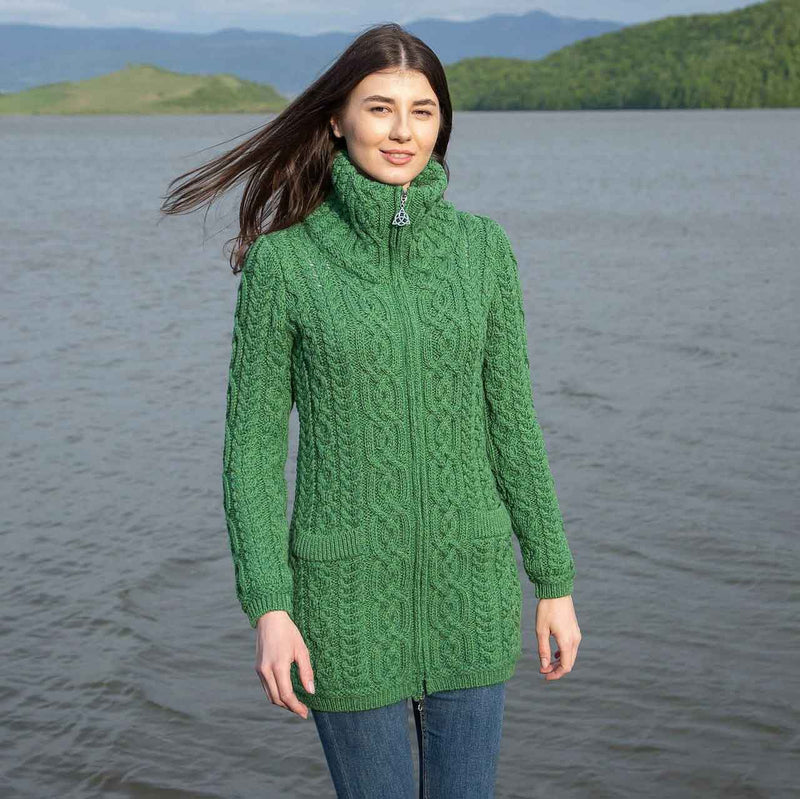 Woman wearing a green Irish soft-merino Aran knit ladies zip cardigan standing by a lake with mountains in the background