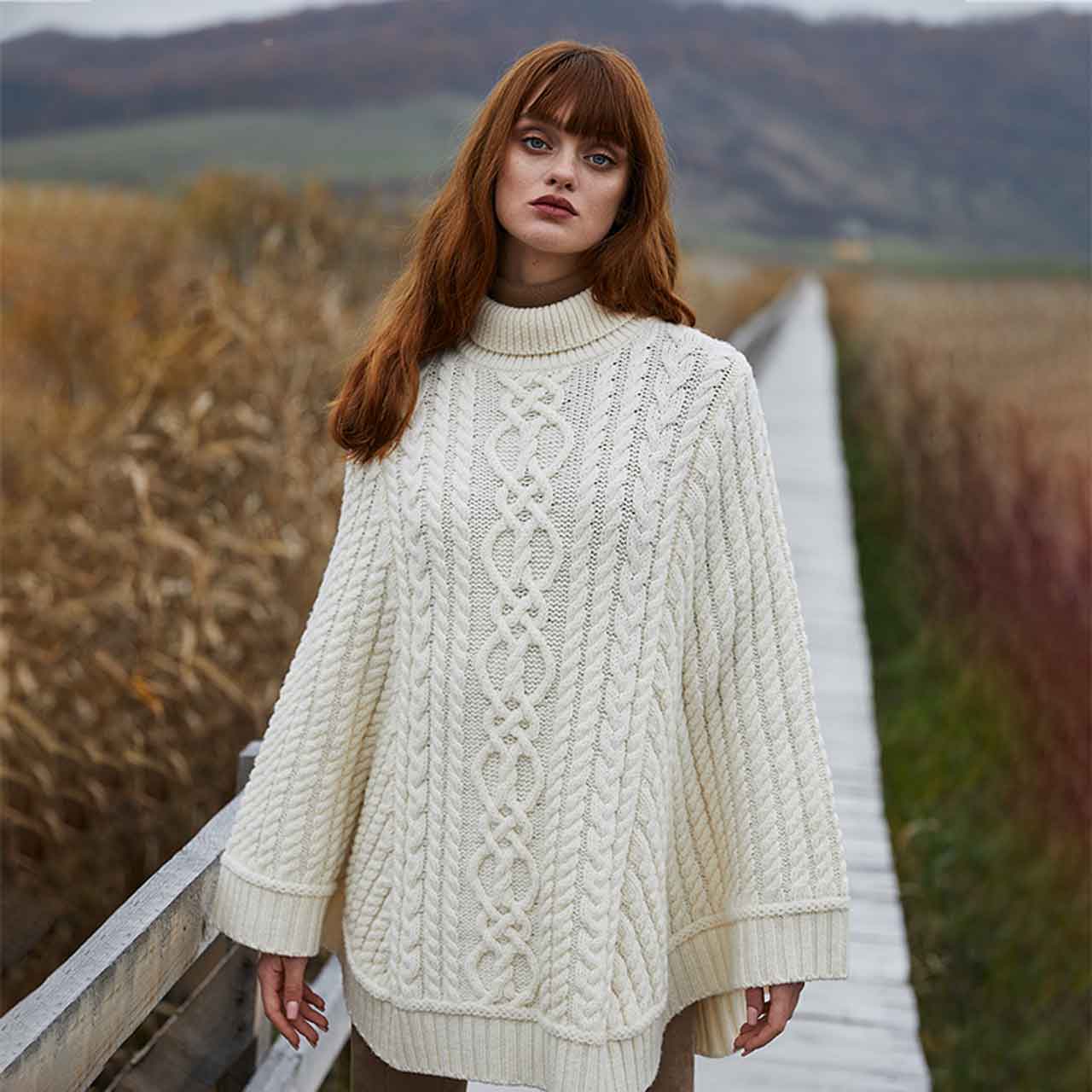 woman wearing a aran cable knit poncho in color natural, standing on a wooden bridge with a scenic background