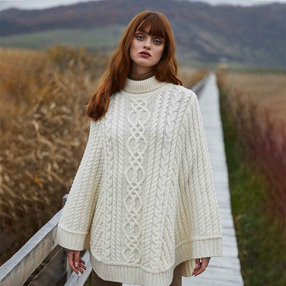 Woman wearing a Aran Cable Knit Poncho in color Natural, standing on a wooden bridge with a scenic background