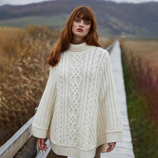 Woman wearing a Aran Cable Knit Poncho in color Natural, standing on a wooden bridge with a scenic background