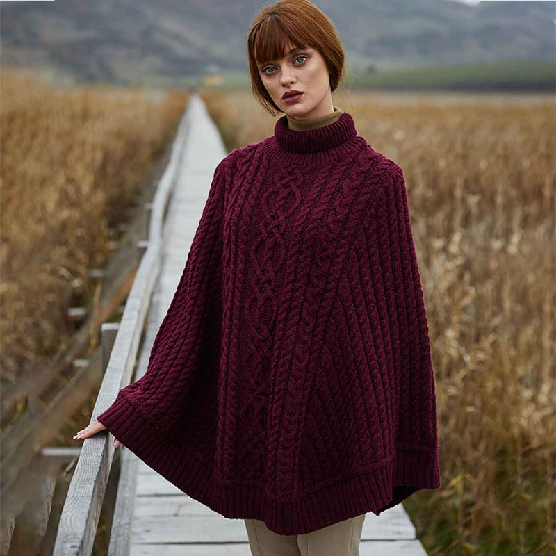 Woman wearing a wine colored Aran Cable Knit Poncho standing on a wooden bridge with a natural landscape in the background