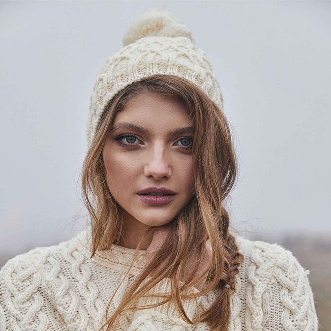 woman wearing a natural/white-colored aran knit merino wool lined ladies beanie and sweater against a neutral background