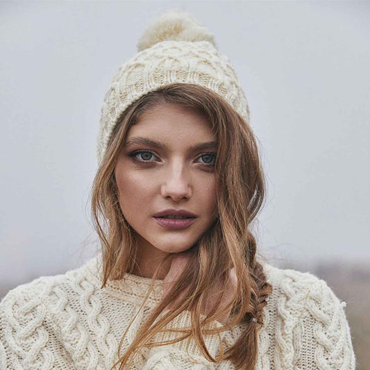 Woman wearing a natural/white-colored aran knit merino wool lined ladies beanie and sweater against a neutral background