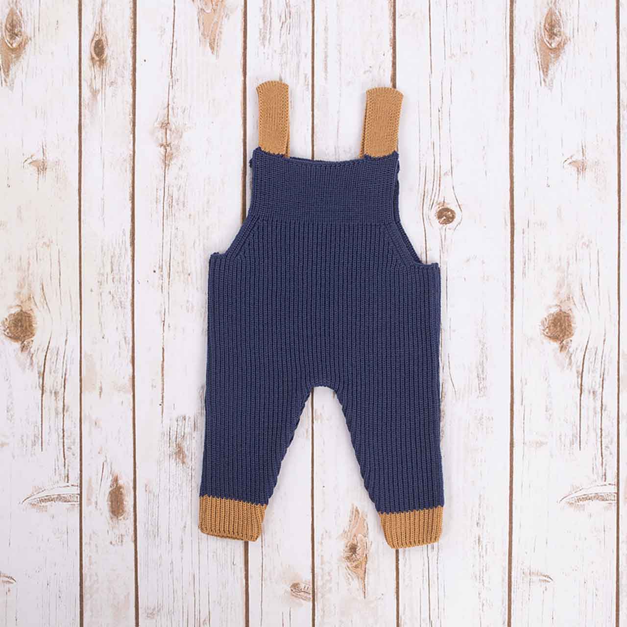 reverse view of navy blue irish baby knitted romper overalls with brown cuffs and buttons on a wooden background