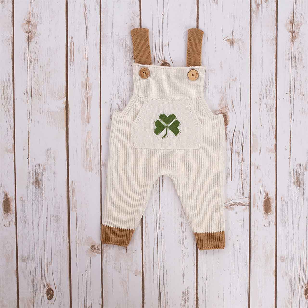 white irish baby knitted romper overalls with a green shamrock knitted pocket on the front, brown cuffs and buttons on a wooden background