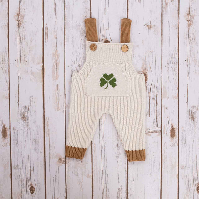 White Irish baby knitted romper overalls with a green shamrock knitted pocket on the front, brown cuffs and buttons on a wooden background