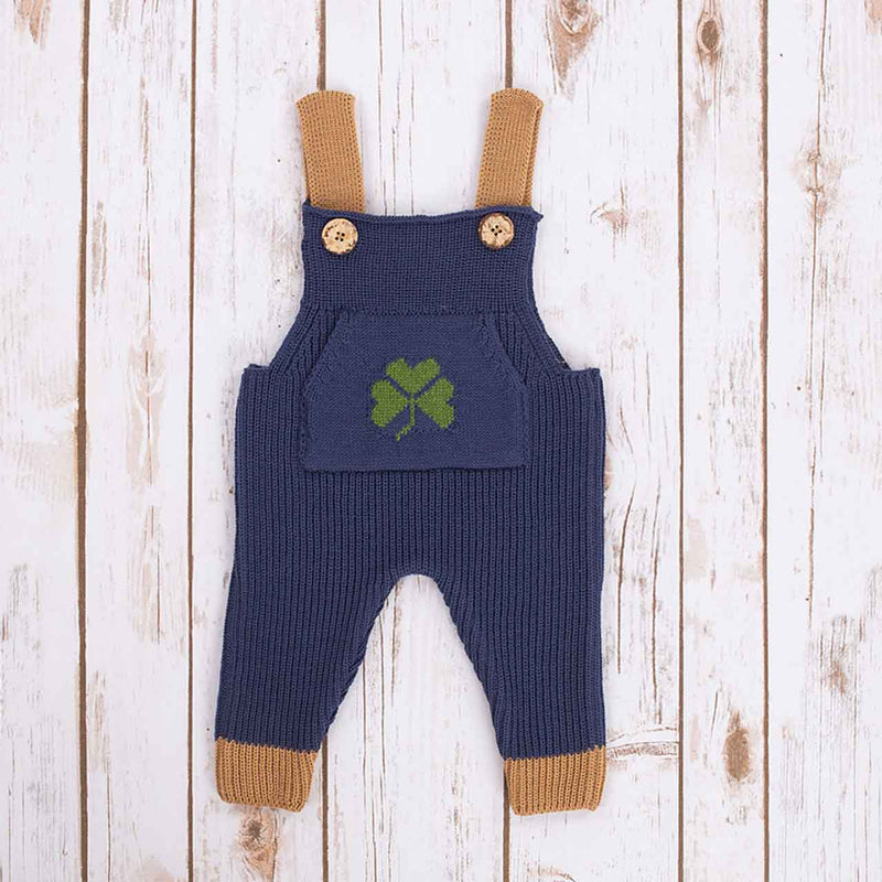 Navy Blue Irish baby knitted romper overalls with a green shamrock knitted pocket on the front, brown cuffs and buttons on a wooden background