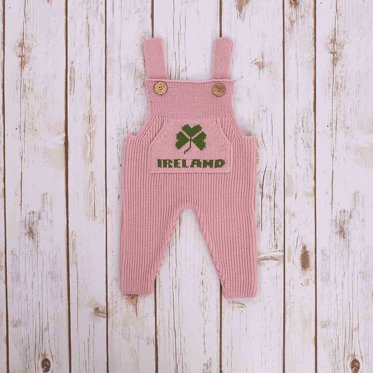 pink irish baby knitted romper overalls with a green shamrock and ireland knitted pocket on the front, and buttons on a wooden background