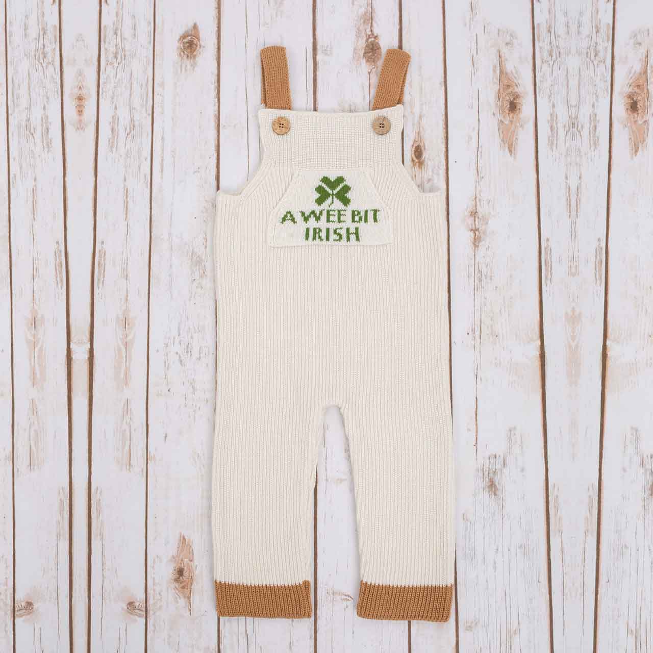white irish baby knitted romper overalls with a green shamrock and a wee bit irish text knitted pocket on the front, brown cuffs, and buttons on a wooden background