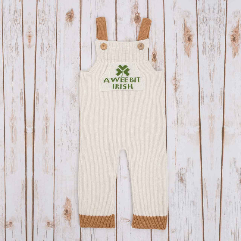 White Irish baby knitted romper overalls with a green shamrock and A wee bit Irish text knitted pocket on the front, brown cuffs, and buttons on a wooden background