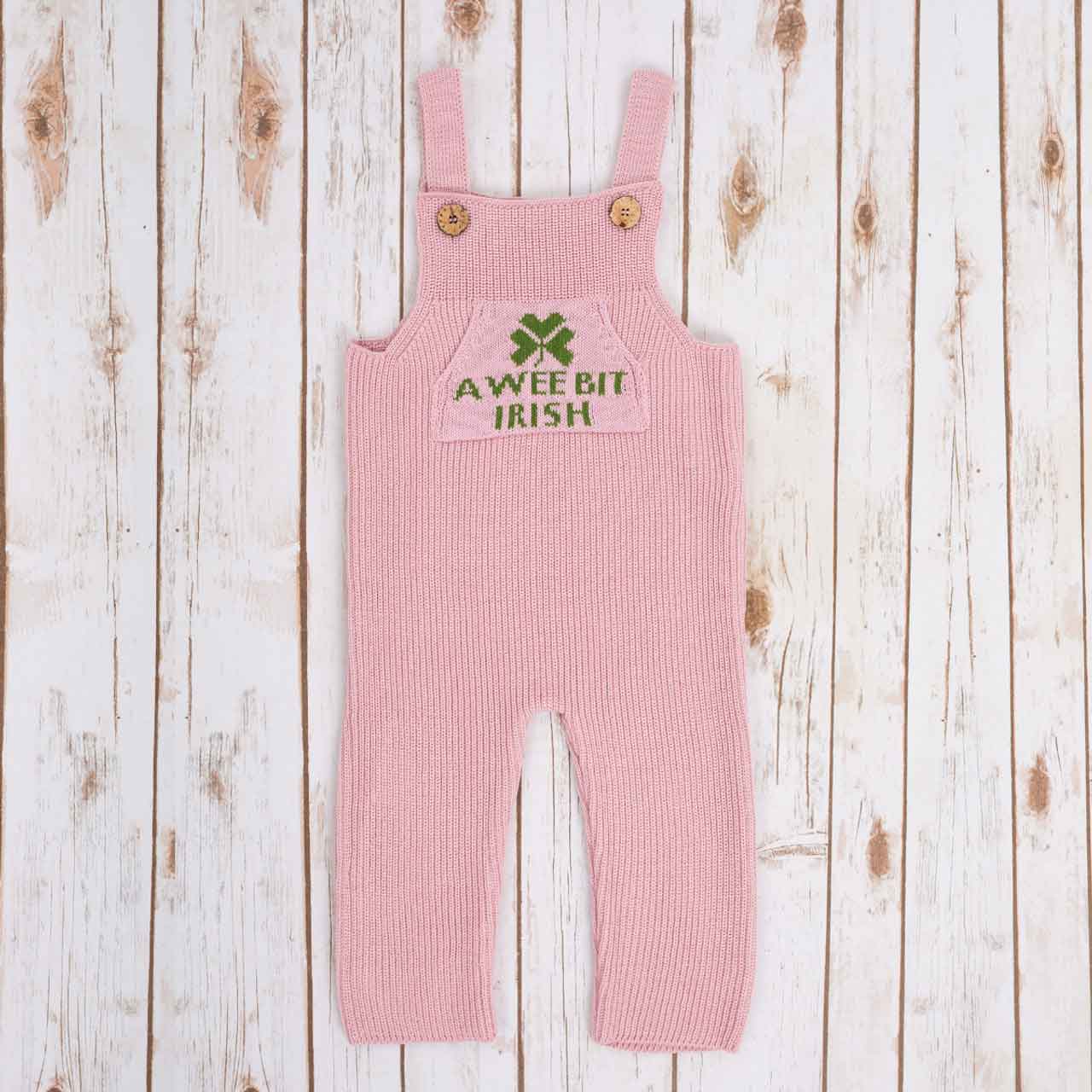 pink irish baby knitted romper overalls with a green shamrock and 'a wee bit irish' text knitted pocket on the front, and buttons on a wooden background