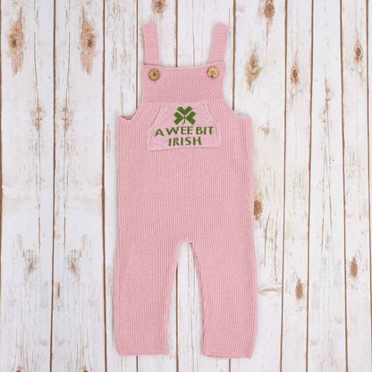 Pink Irish baby knitted romper overalls with a green shamrock and 'A wee bit Irish' text knitted pocket on the front, and buttons on a wooden background