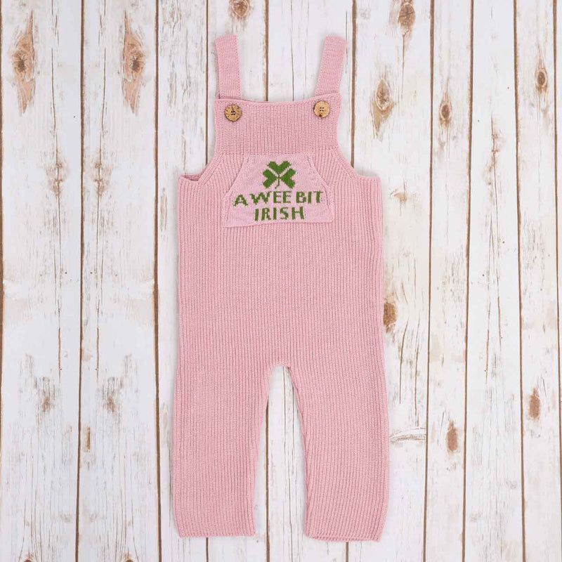 Pink Irish baby knitted romper overalls with a green shamrock and 'A wee bit Irish' text knitted pocket on the front, and buttons on a wooden background