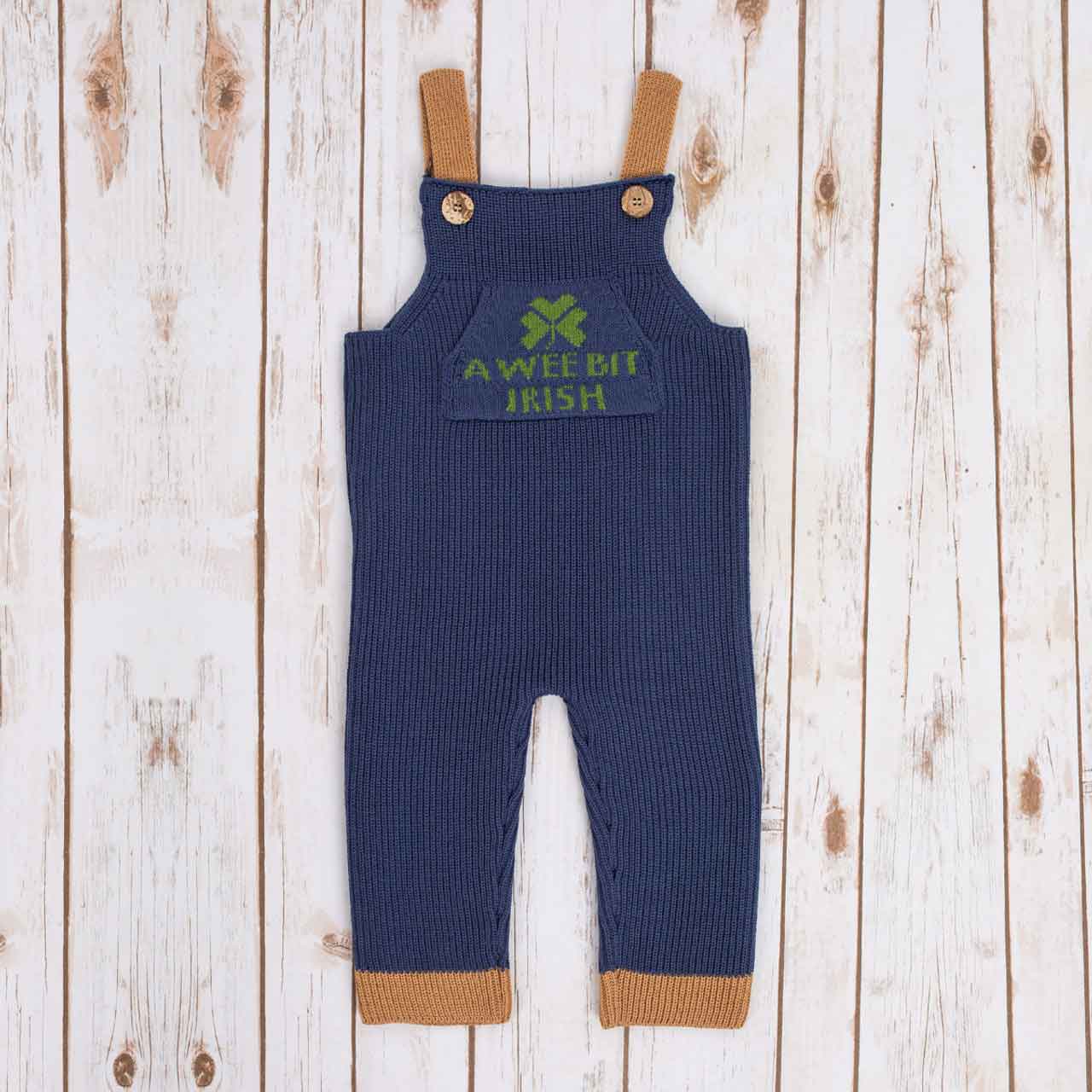 navy blue irish baby knitted romper overalls with a green shamrock and 'a wee bit irish' text knitted pocket on the front, brown cuffs, and buttons on a wooden background