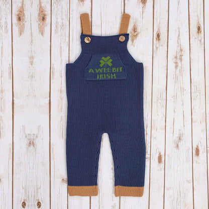 Navy Blue Irish baby knitted romper overalls with a green shamrock and 'a wee bit Irish' text knitted pocket on the front, brown cuffs, and buttons on a wooden background