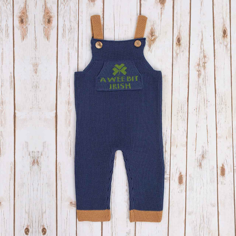 Navy Blue Irish baby knitted romper overalls with a green shamrock and 'a wee bit Irish' text knitted pocket on the front, brown cuffs, and buttons on a wooden background