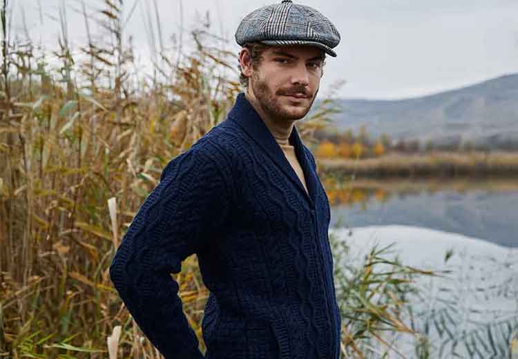 Man wearing a navy Mens Aran Knit Zipper Cardigan  and flat cap standing by a lake with mountains in the background.
