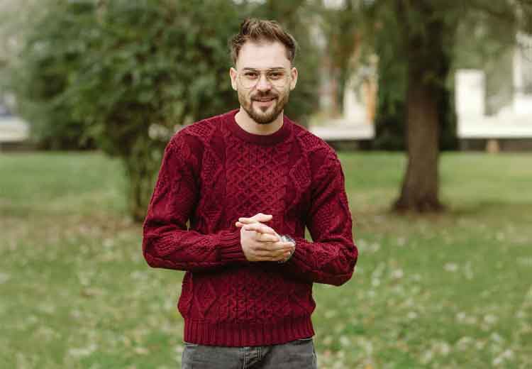 Man wearing a Wine Irish Fisherman Cable Knit Men's Sweater standing outdoors with trees and grass in the background