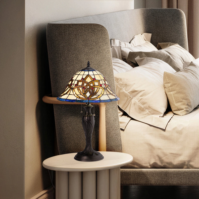 A Celtic Blooming Tiffany-style stained glass table lamp with an ornate base sits on a small round side table next to a bed with beige bedding and a gray upholstered headboard.