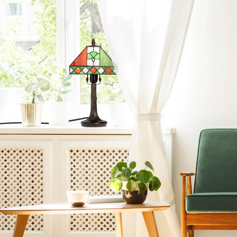A St. Patrick's Day Irish Flag Tricolor Stained Glass Table Lamp sits on a windowsill in a bright room with white curtains, a small wooden table with a potted plant, and a green armchair.