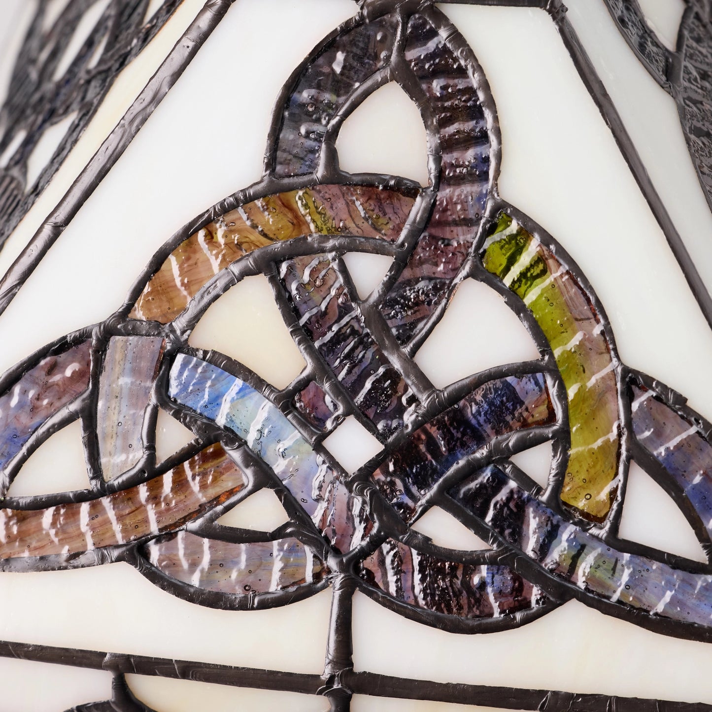close-up of a celtic trinity knot stained glass table lamp featuring a celtic triquetra knot design with iridescent segments in purple, blue, green, and brown, held together by dark metal solder over a cream glass background.
