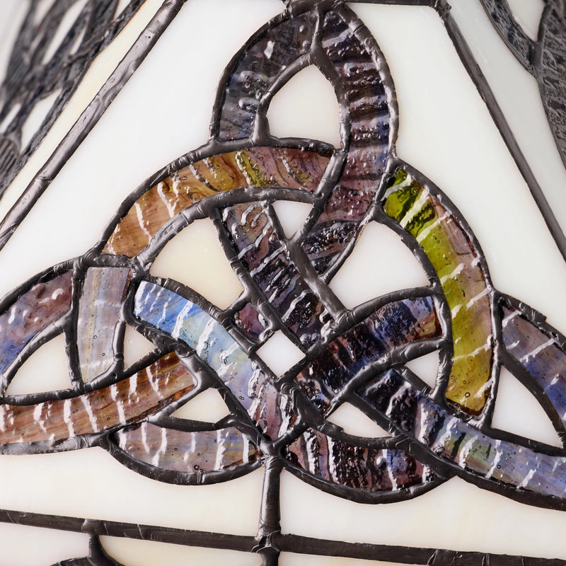 Close-up of a Celtic Trinity Knot Stained Glass Table Lamp featuring a Celtic triquetra knot design with iridescent segments in purple, blue, green, and brown, held together by dark metal solder over a cream glass background.
