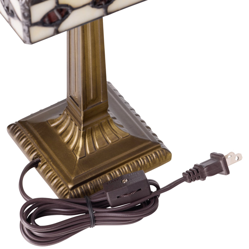 A close-up of a table lamp's base and power cord on a white background. The lamp has a square, ornate bronze-colored base and a stained-glass shade with cream, black, and brown mosaic patterns.