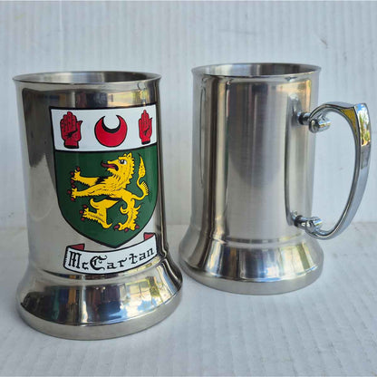 Front and reverse views of a stainless steel Coat of Arms Hand Painted Tankard, hand painted with the McCartan coat of arms on a grey surface. 