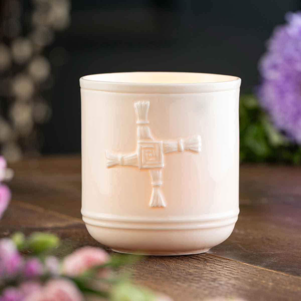 belleek pottery st. bridget's cross irish votive jar on a wooden surface with blurred flowers in the background